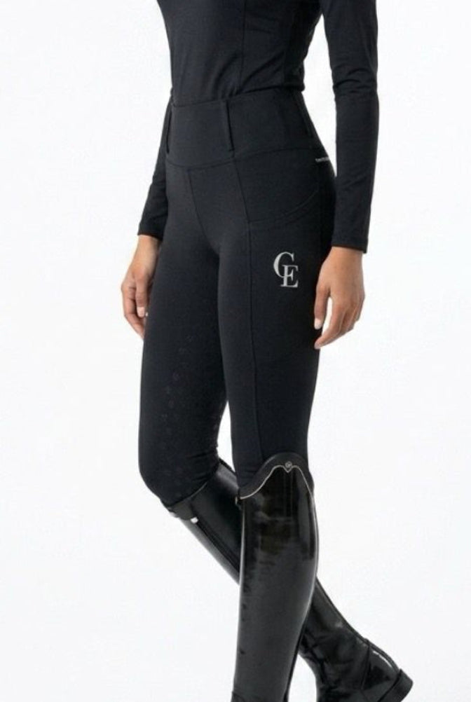Pantalón de equitación negro ajustado con logo CE bordado en el muslo, combinado con base layer negro de manga larga, llevado por una amazona con botas de campo negras, vista lateral, fondo blanco