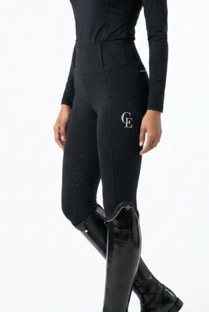 Pantalón de equitación negro ajustado con logo CE bordado en el muslo, combinado con base layer negro de manga larga, llevado por una amazona con botas de campo negras, vista lateral, fondo blanco
