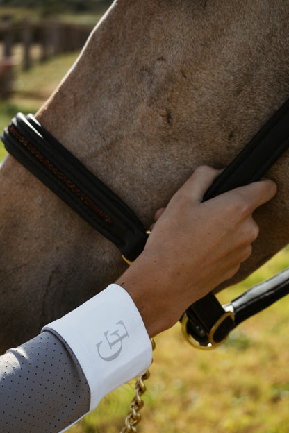 Mano de amazona con camisa de concurso gris perforada Celiana Equestrian, puño blanco con logo bordado, apoyada en la cabeza de un caballo con cabezada de cuero negro. 