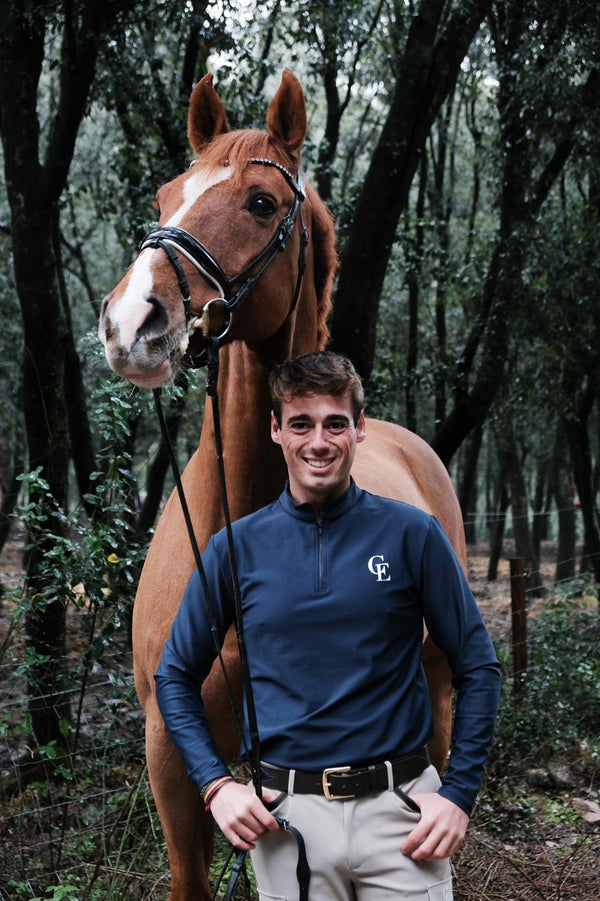 Jinete hombre con polo técnico azul marino Celiana Equestrian con logo CE, posando junto a su caballo castaño en entorno natural.