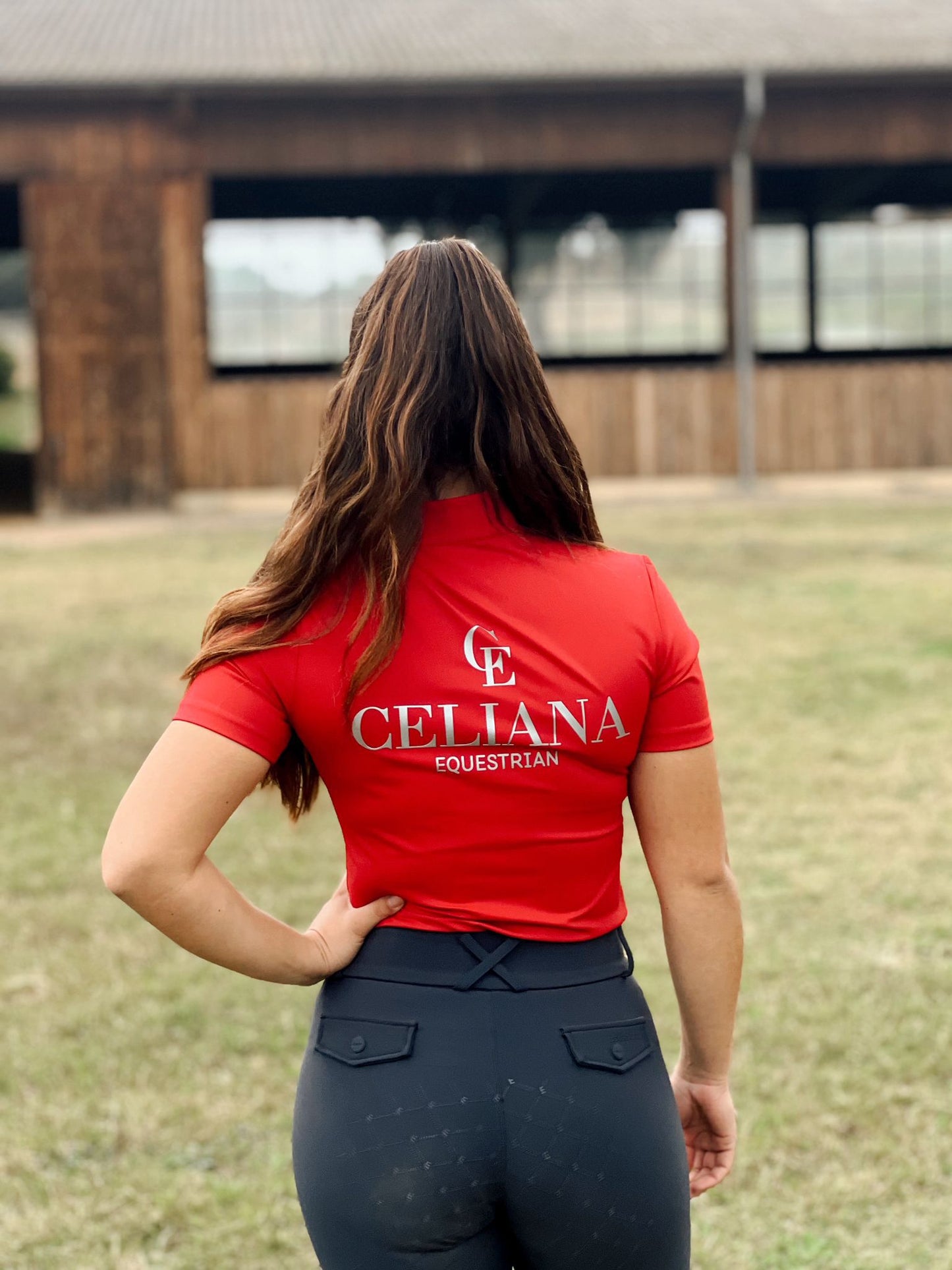 Amazona de espaldas con base layer roja de equitación Celiana Equestrian Prestige de manga corta con logo CE y ajuste técnico elegante.