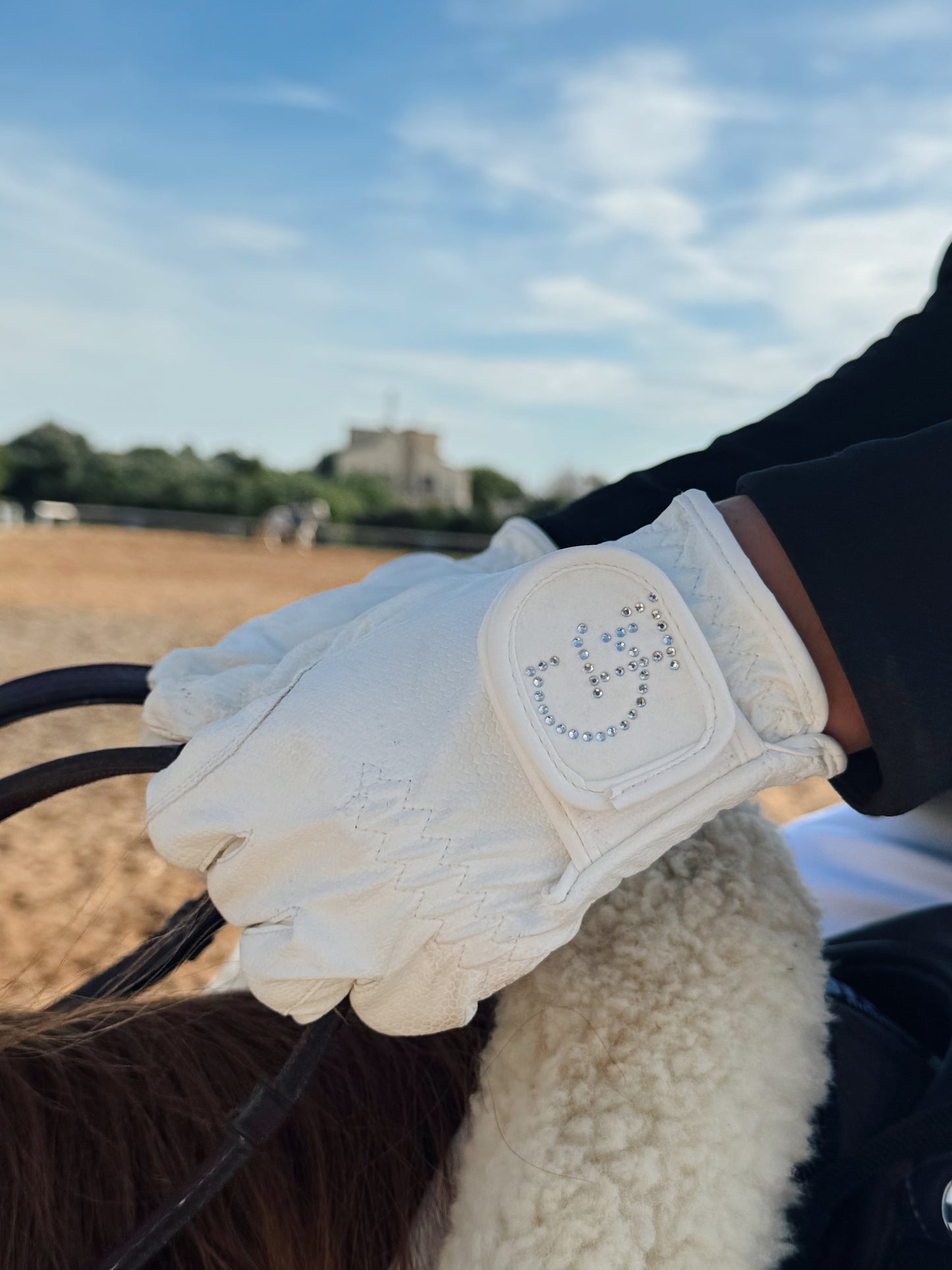 Guante técnico de equitación blanco Celiana Equestrian con logo CE en cristales, amazona sujetando riendas sobre caballo, al aire libre