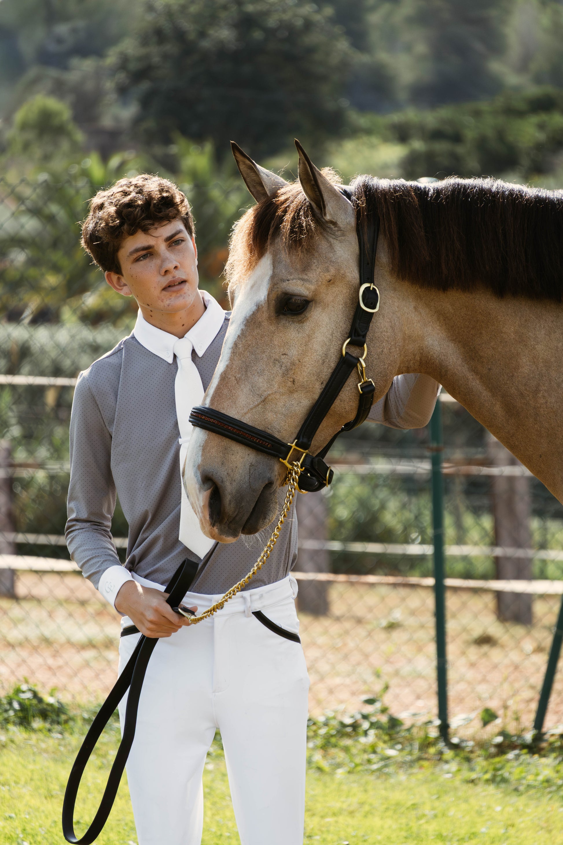 Jinete joven con pantalón de equitación blanco Celiana Equestrian y camisa gris, junto a caballo bayo con cabezada negra, en exterior