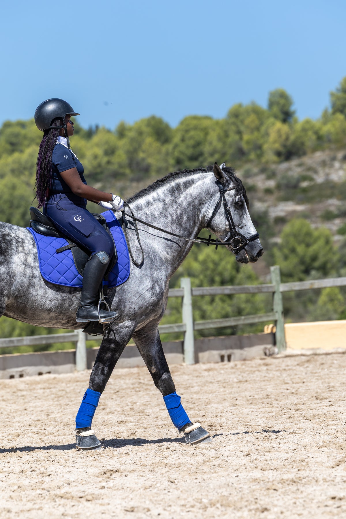 Amazona al paso sobre un caballo tordo en pista de doma, con vendas de trabajo y sudadero azules Celiana Equestrian