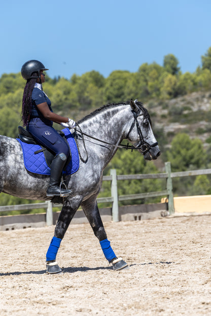 Amazona al paso sobre un caballo tordo en pista de doma, con vendas de trabajo y sudadero azules Celiana Equestrian